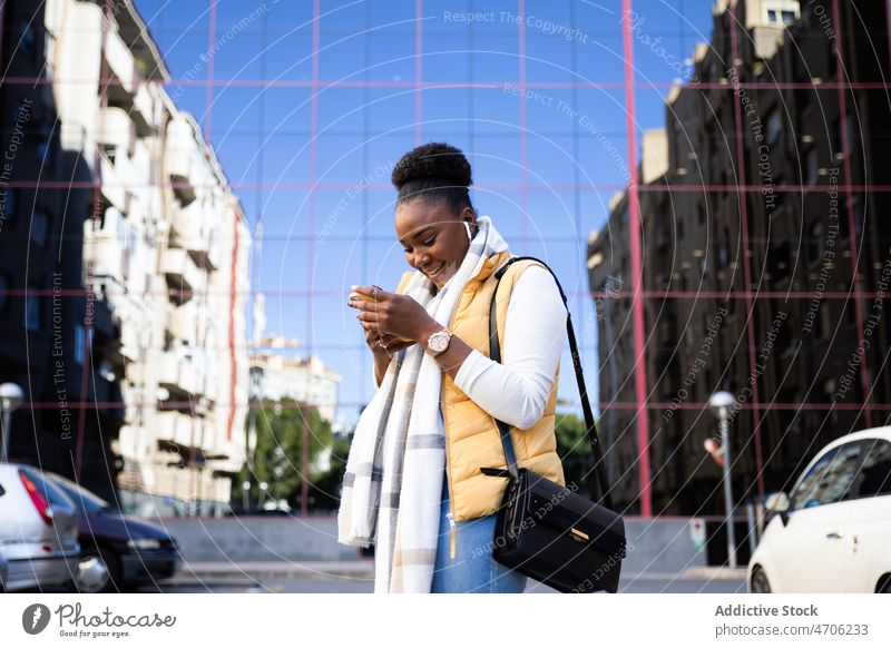 Cheerful black woman browsing phone on street watching smartphone using texting stand looking down conversation discuss earphones communicate city earbuds