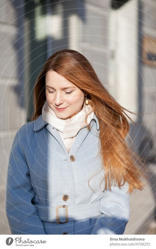 Young beautiful woman in a light blue coat standing on the street in the city in sunlight on sunny day. Candid lifestyle portrait of a woman, looking at the camera and smiling.