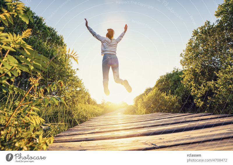 Happy woman jumping with raised hands and legs, and enjoying life over a Wooden path at sunset happy free nature trampoline air girl walkway weekend boardwalk