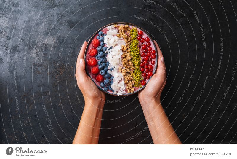 Woman hands hold a Healthy breakfast bowl, wirh blueberry smoothie with, raspberry, blueberrys, coconut, nuts and currants toppings healthy fruit dessert