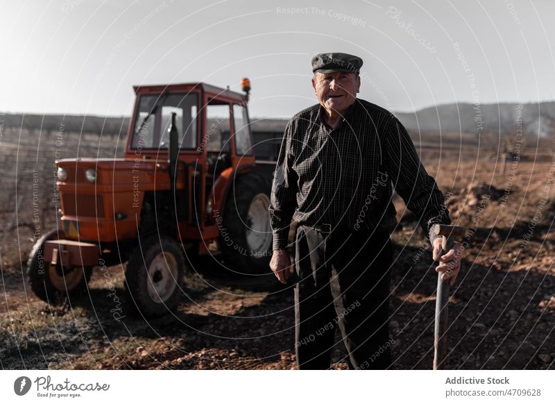 Smiling senior man with gardening tool near tractor in field farmer agriculture cart rural work countryside male transport agronomy positive plantation job