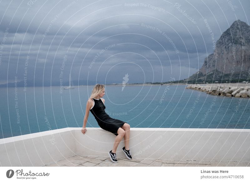 A picturesque though a bit moody background behind a gorgeous woman. She is a muse. Dressed in a black dress and wearing sneakers casually. Dark blue clouds are gathering. A beautiful coastline of San Vito Lo Capo in Sicily.