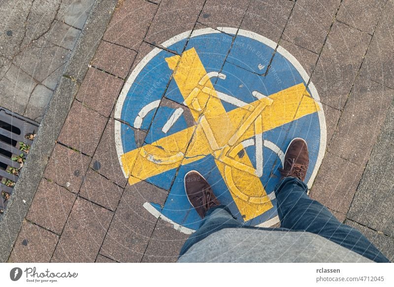 young man stands on a Bicycle path with cross sign at a sidewalk, high angle footsie or flortrait, personal pespective from above. pov people lifestyle point