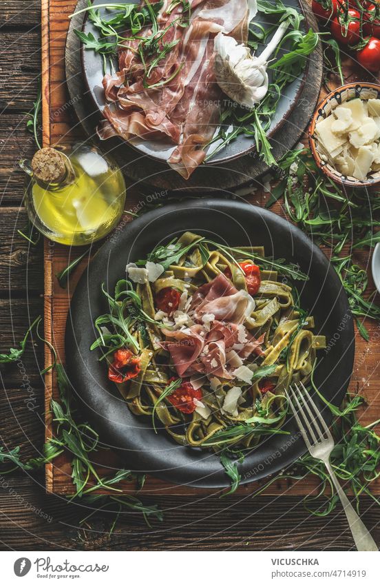 Green tagliatelle pasta with ham, parmesan cheese, arugula and tomatoes on black plate green rustic wooden kitchen table ingredients olive oil cutlery top view
