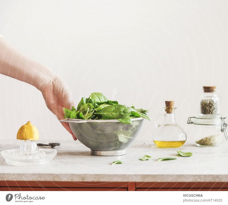 Woman hand holding metal sieve filled with raw green spinach leaves on kitchen table woman lemon olive oil herbal salt pepper jars white wall background cooking