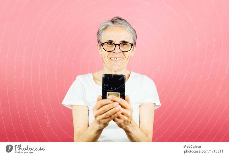 Old woman checking his smart phone happy while smiling, giving an warning posing isolated over pink color wall background. Daily expressions with copy space in studio.