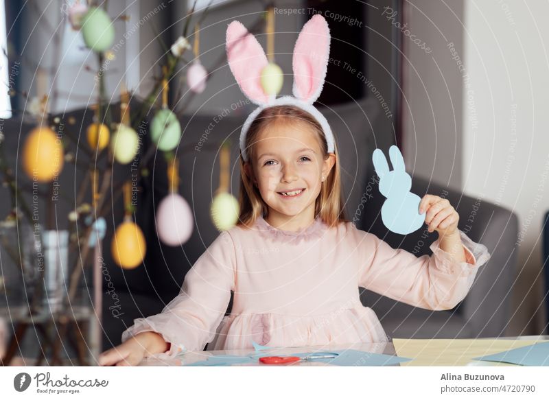 happy caucasian toddler girl eight years old at home in living room making garland with paper bunny. Stay home during Coronavirus covid-19 pandemic easter egg