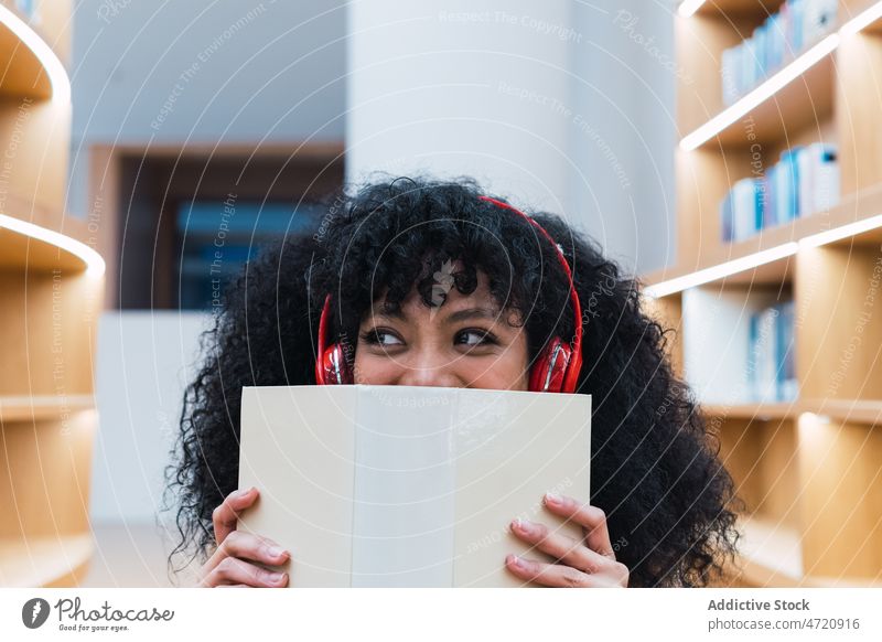 Positive ethnic lady in headphones with opened book in library woman meloman music dreamy listen interesting literature female shelf bookshelf bookcase positive