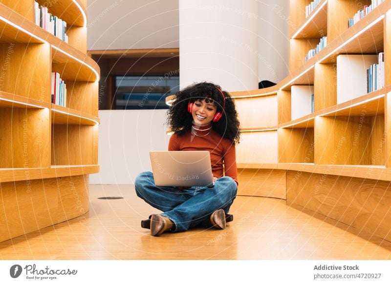 African American lady using laptop in library to music woman meloman student headphones smile bookshelf bookcase listen browsing education enjoy ethnic happy