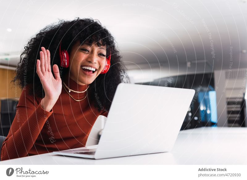 Ethnic female with laptop showing greeting gesture during video call woman hello wave hand hi using headphones smile browsing enjoy ethnic happy