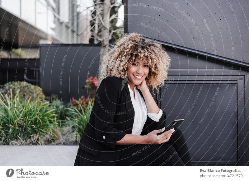 beautiful smiling business woman using mobile sitting on stairs in city. Buildings background afro hispanic mobile phone skyscraper building young curly hair