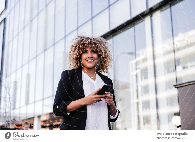 beautiful smiling business woman using mobile phone and headphones in city. Buildings background afro hispanic skyscraper building young curly hair music