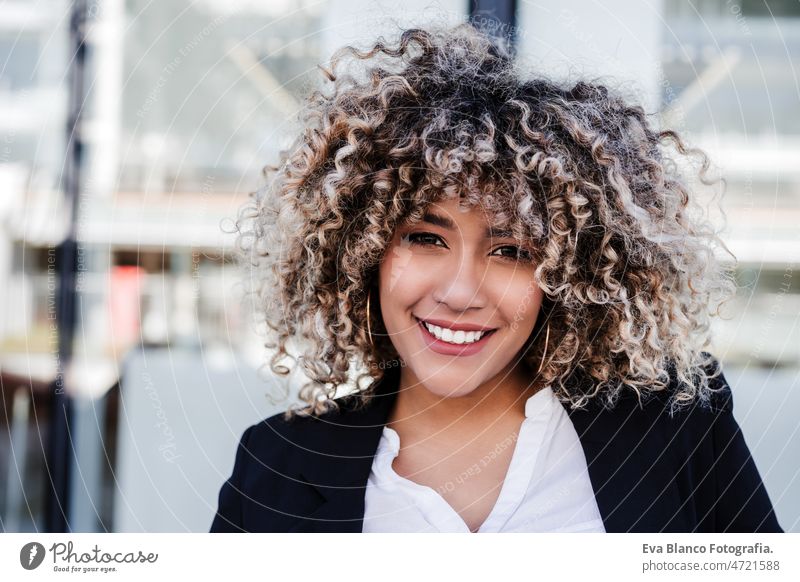 portrait of confident hispanic business woman in city. Buildings background afro mobile phone skyscraper building young curly hair beautiful headphones music