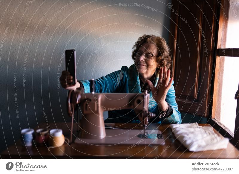 Cheerful seamstress taking selfie near sewing machine woman handicraft smartphone wave hand hobby mature senior pensioner appliance thread instrument female