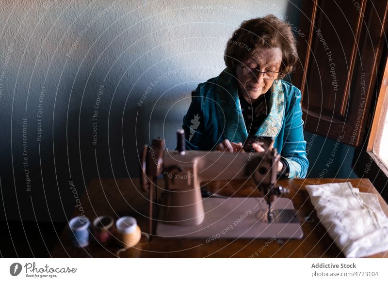 Senior woman sewing on machine sewing machine textile seamstress handicraft hobby appliance thread instrument female style yarn design room craftswoman table