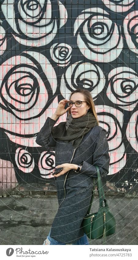 A young woman in a gray coat, green scarf adjusts glasses against the background of a colored wall model fashion bag beautiful style girl pretty street stylish