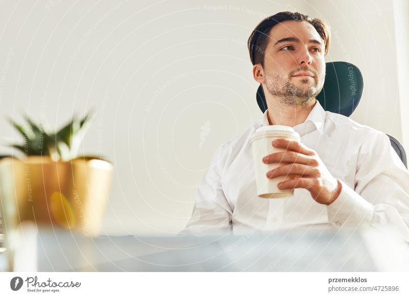 Young confident businessman sitting at desk in office. Relaxed man wearing white shirt holding cup of coffee looking away working entrepreneur colleague manager