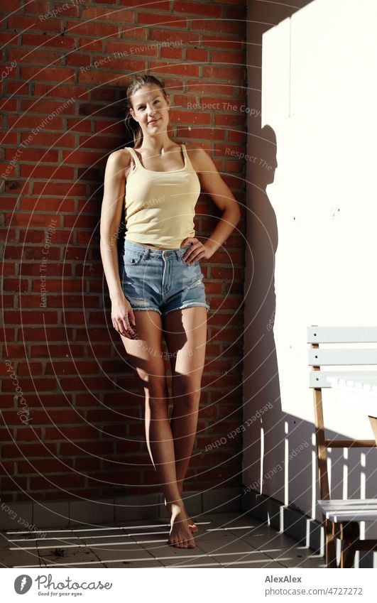 Young tall blonde woman standing on balcony barefoot in the sun leaning against brick wall and smiling at camera Woman pretty Large Slim Esthetic Athletic fit