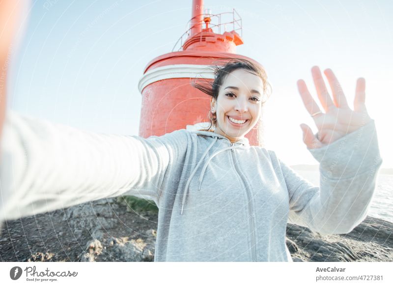 Young arab woman taking a selfie on the seashore lighthouse during a trip travel while smiling to camera. Happy experiences while road trip, exploring new lands and taking photos for the family