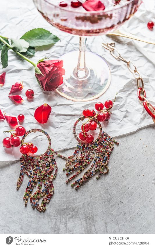 Close up of beautiful earrings on light table with red currants, rose flower and champagne glass close up grey concrete jewelry drink concept top view