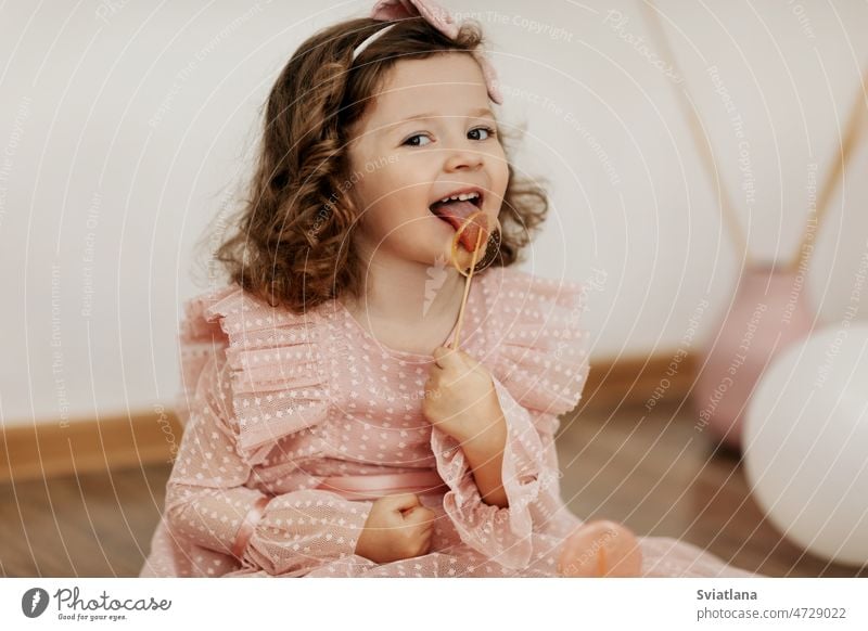 A little girl in a pink dress is sitting on the floor and eating a lollipop child candy cute sweet children and sugar baby caucasian adorable pretty portrait