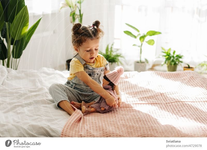 A little girl is sitting on the bed in the bedroom and playing with her doll, tying her scarf kid toy childhood beautiful caucasian baby background face cute