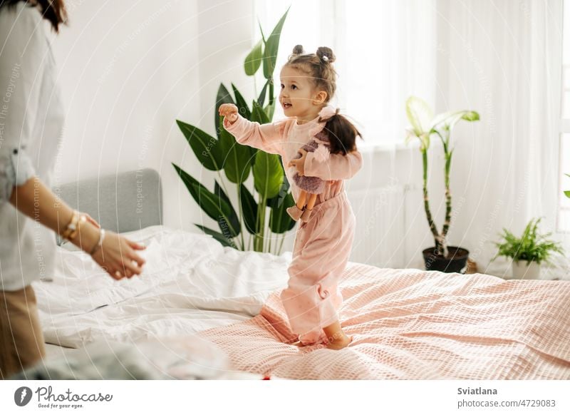 A charming little girl plays with dolls with her mom on the bed during the day. Motherhood, caring, time together daughter pretty portrait family happy mother