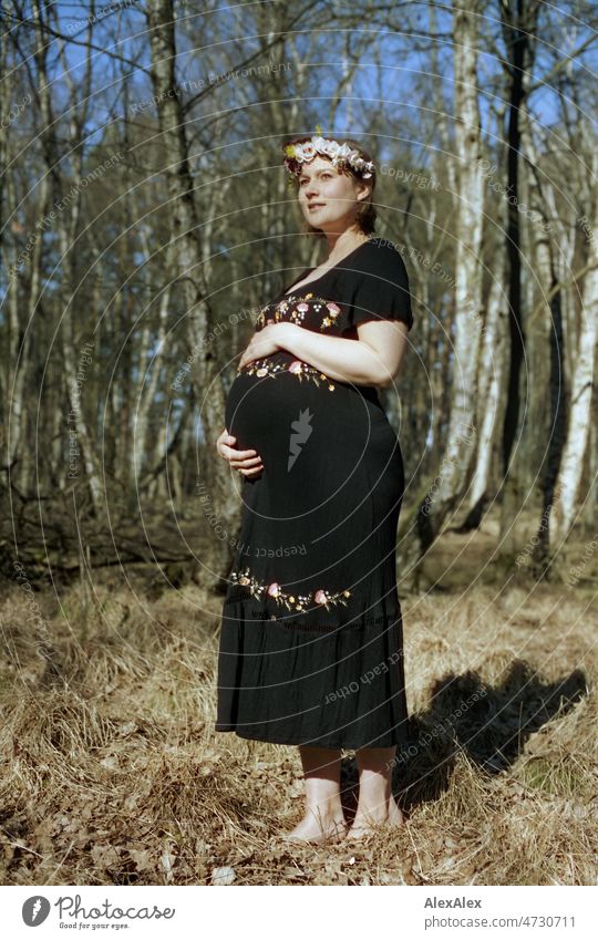 Analog full body portrait of young pregnant woman standing barefoot in a deciduous forest with wreath of flowers in her hair and holding her baby bump with one hand