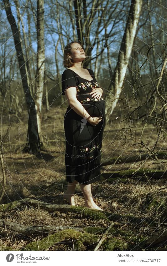 Analog full body portrait of young pregnant woman standing barefoot in a deciduous forest holding her baby belly with one hand Smiling vigorous Grass Woodground