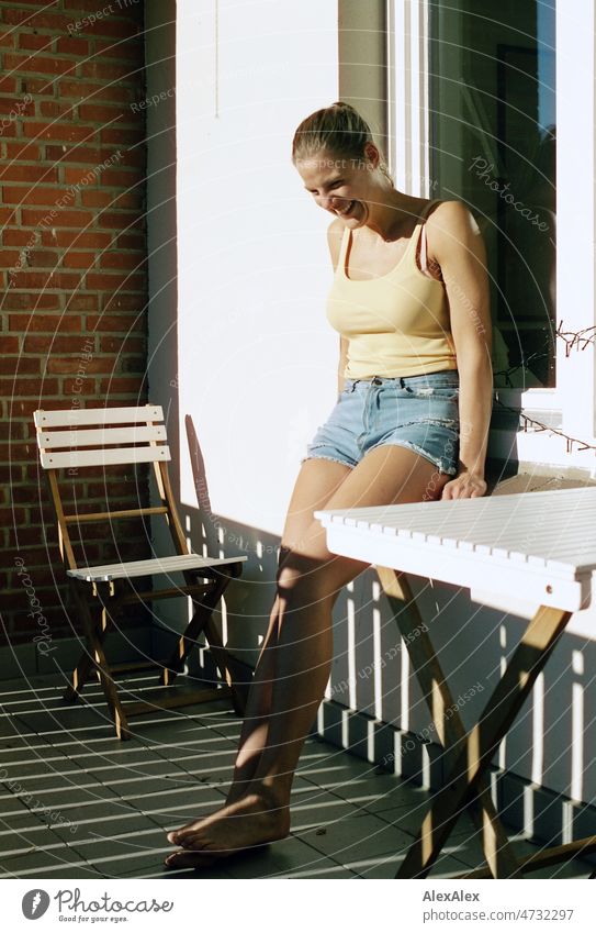 Young tall blonde woman leaning against window on balcony barefoot in the sun and smiling - analog portrait Woman pretty Large Slim Esthetic Athletic fit