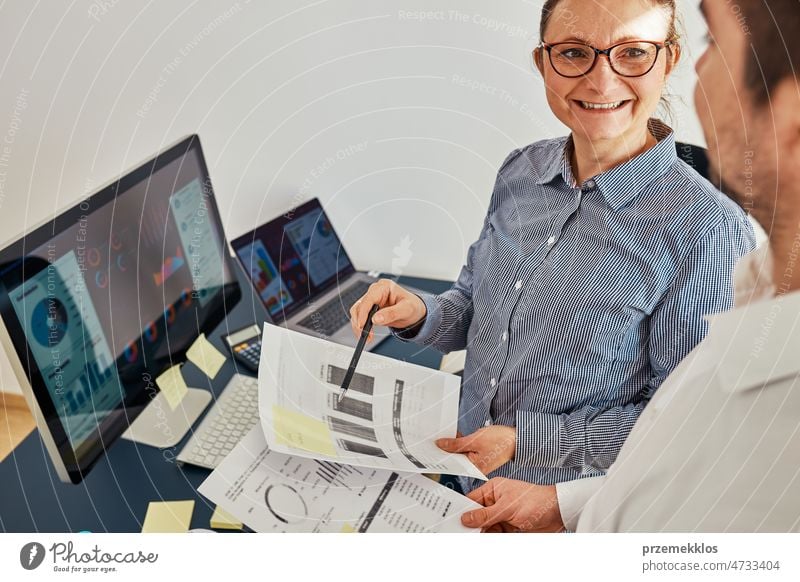 Woman entrepreneur discussing financial data with her colleague standing at desk in office. Happy positive businesswoman working with charts and tables on computer. Two people working together