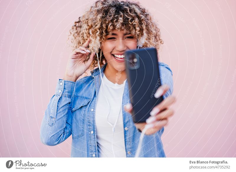 portrait of smiling hispanic woman with afro hair in city using mobile phone and headset. lifestyle curvy body positivity selfie technology internet wireless