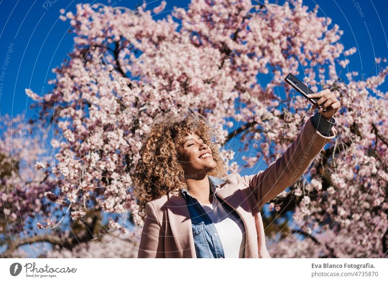 smiling hispanic woman in park taking picture with mobile phone. Spring flowers background spring technology smart phone internet device dancing curvy