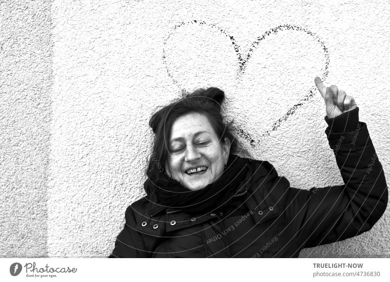 Happily relaxed, eyes closed, the laughing woman, warmly dressed and her long dark hair pinned up, points to the large heart sign behind her on the white wall.