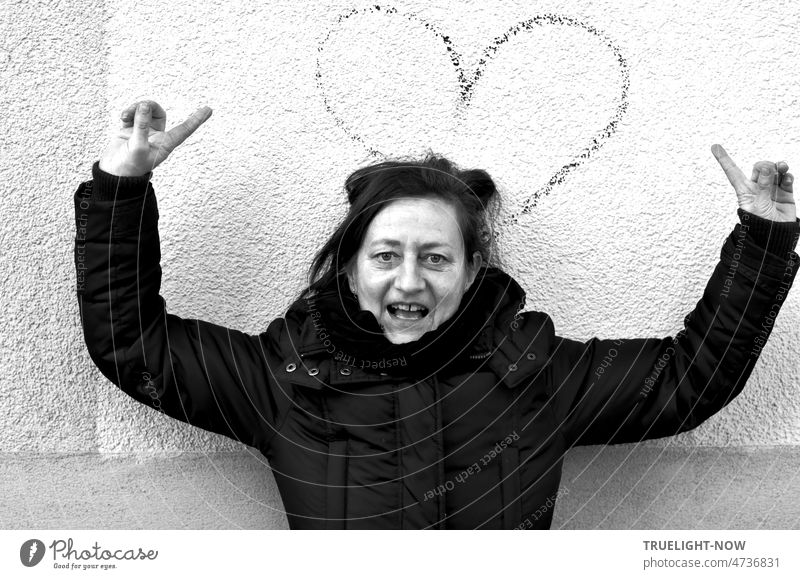 "Listen to your heart!" the woman shouts to the photographer, dramatically raising her arms and hands in an imploring manner, her index fingers pointing energetically at the heart sign behind her on the white wall