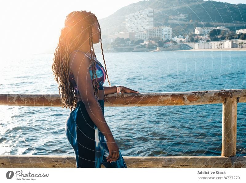 Black woman in sportswear leaning on fence on embankment fit sea training thoughtful wellness healthy activewear female sporty serious confident slim waterfront