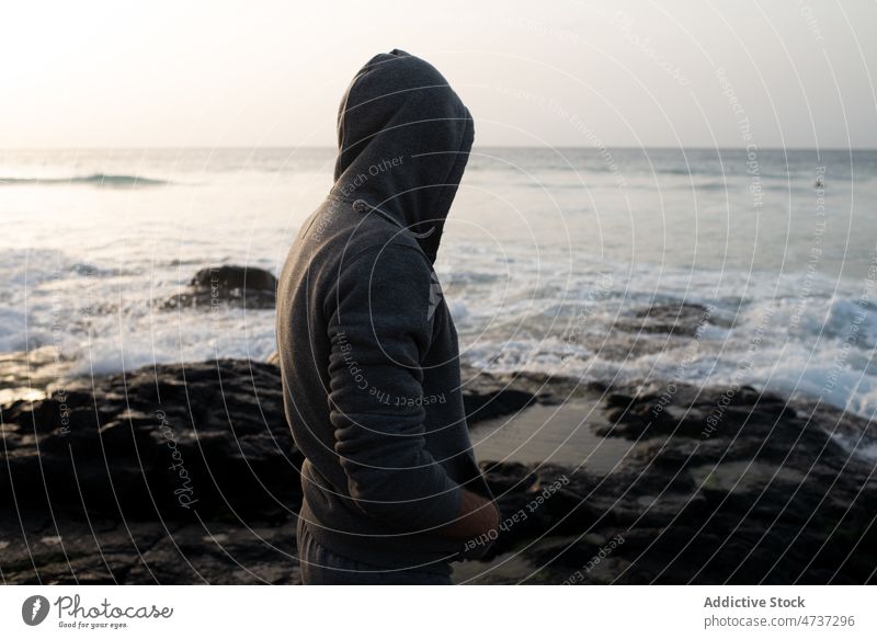 Anonymous man in hood standing on beach admire ocean vacation contemplate harmony freedom serene trip male sand island shore coast seashore hoodie nature adult