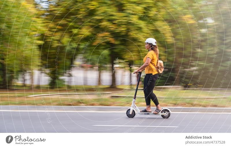 Smiling black woman riding scooter in alley ride helmet park protect eco friendly activity electric female path walkway vehicle tree pathway hobby safety plant