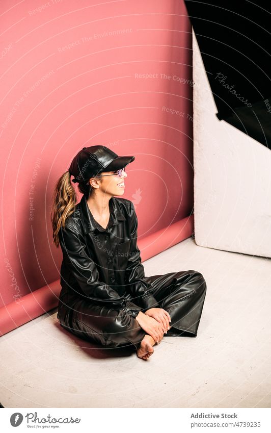 Female model on floor during photo session woman studio posing posture take shoot leather jacket cap trendy style cheerful outfit smile female slim positive