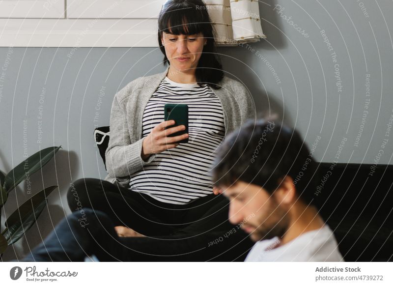 Pregnant wife browsing smartphone in apartment near husband couple digital addict using connection internet read living room man woman pregnant device