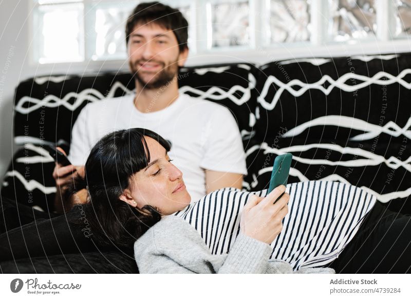 Young Hispanic man watching TV on sofa near pregnant wife using smartphone couple rest tv spend time lying relationship free time relax switch remote control
