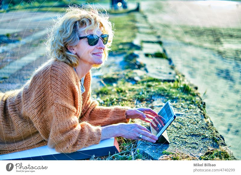 Young mature blonde caucasian woman relaxing outdoor with a tablet in a sunny day. lifestyle portrait women healthy connected technology internet wireless