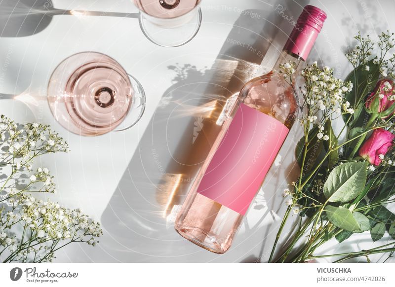 Rose wine bottle with empty pink mock up label, wineglasses and flower bouquet sunlight rose wine two wine glasses romantic white background shadow modern