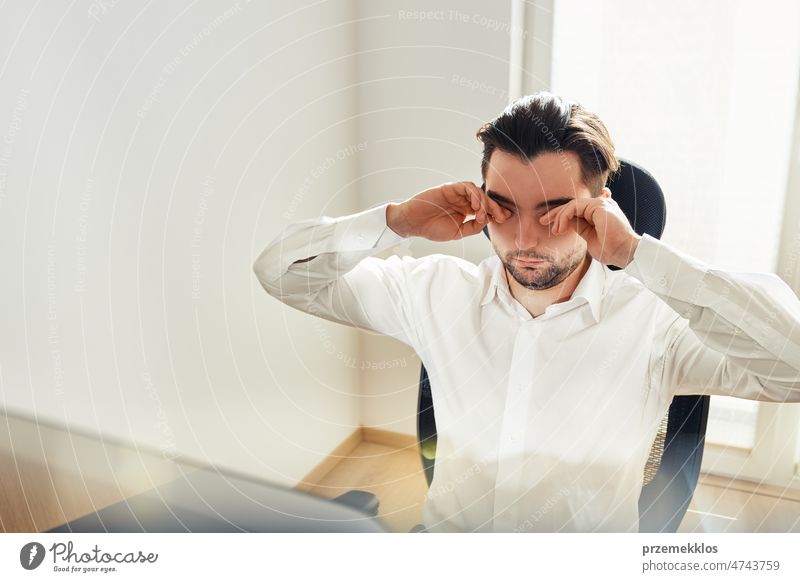 Tired exhausted young man rubbing eyes feeling eyestrain from long computer work. Man sitting at computer screen office working businessman tired sleepy