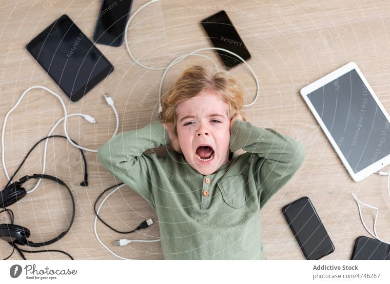 Screaming boy lying amidst gadgets scream upset cable dependency addiction punishment unhappy prohibit modern problem device yell kid smartphone tablet