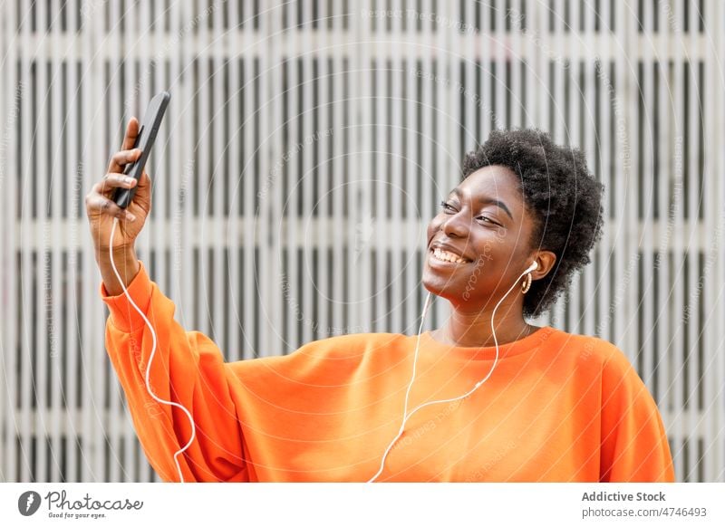Cheerful black woman listening to music while taking selfie earphones meloman song street smartphone social media self portrait capture memory hobby cellphone