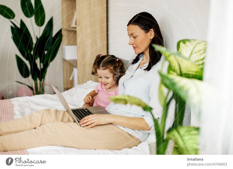 A young mother is working on a laptop or watching videos with her little daughter in the bedroom. Time together, work online family freelance business