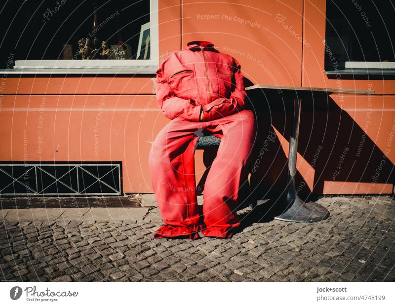 Headless in red overalls he sits at the crooked bistro table Overalls Red Seat Prenzlauer Berg Berlin Torso House wall Doll Cobblestones Clothing without head