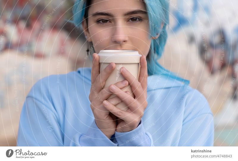 Blue haired Teenage girl in light blue oversize hoodie staying near graffiti wall with coffee to go Teenager mockup cup jeans blue haired teen girl outdoors