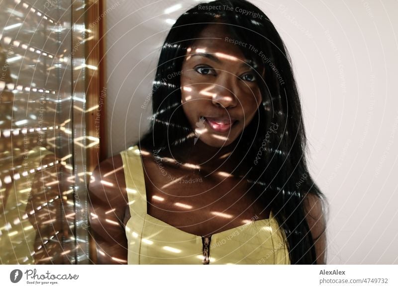Young beautiful long haired woman in yellow summer dress stands in front of half closed blinds, light streaks on her face Woman Young woman pretty